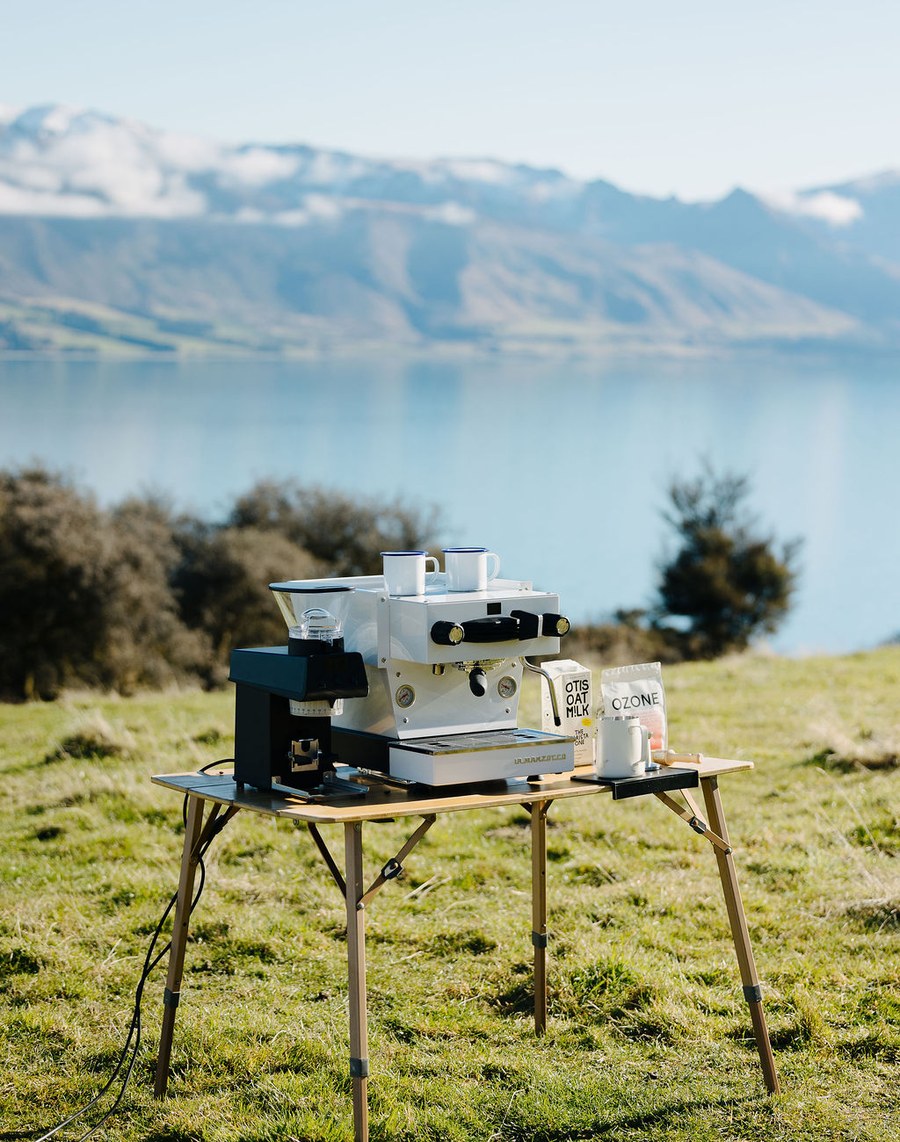 A La Marzocco espresso machine, grinder and Otis Oat M!lk set up on a camp table on a grassy hillside above a lake