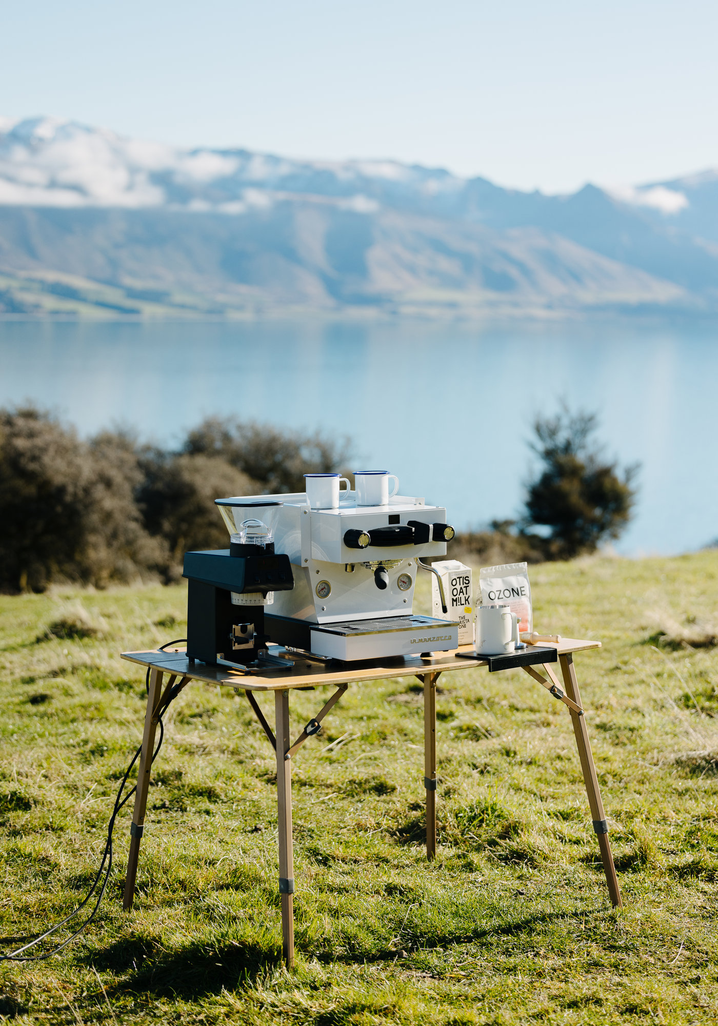 A La Marzocco espresso machine, grinder and Otis Oat M!lk set up on a camp table on a grassy hillside above a lake