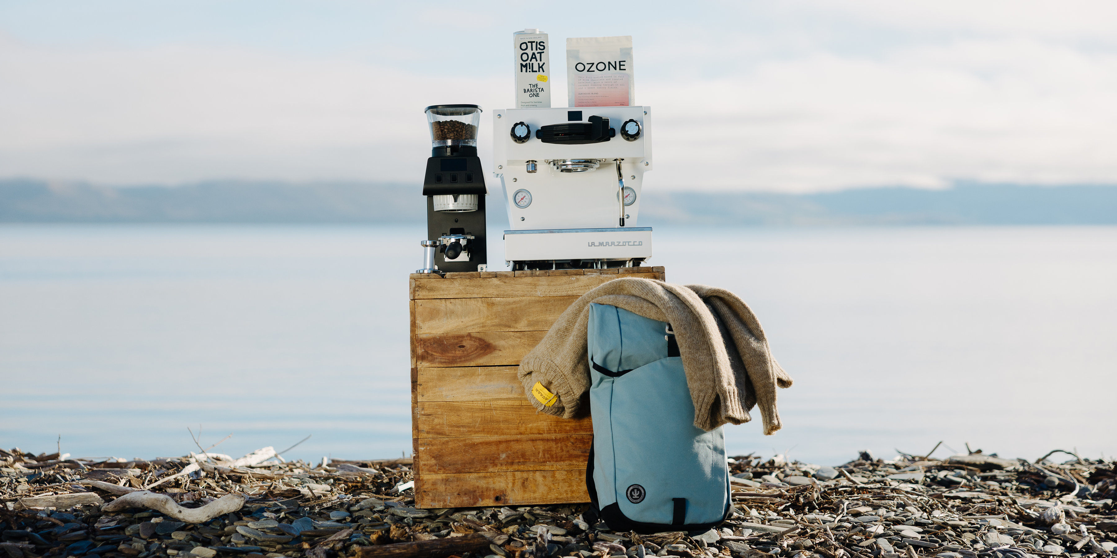 La Marzocco espresso machine, grinder, Otis Oat M!lk and Ozone coffee on a wooden crate beside a lake, with a backpack and knit resting on the stones