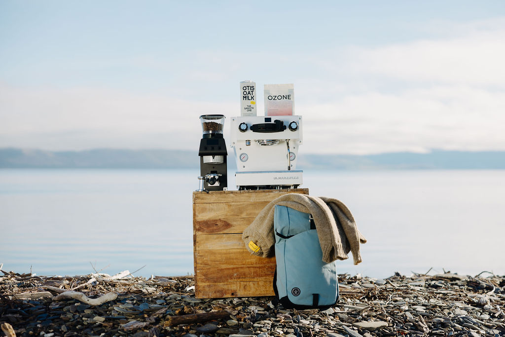 La Marzocco espresso machine, grinder, Otis Oat M!lk and Ozone coffee on a wooden crate beside a lake, with a backpack and knit resting on the stones